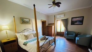 A four-poster bed with a wooden frame, a bedside table, a lamp, a painting on the wall, a wardrobe, a chair, and a ceiling fan. - Spacious Farmhouse Rental in Ladismith With Stunning Views (Ladismith)