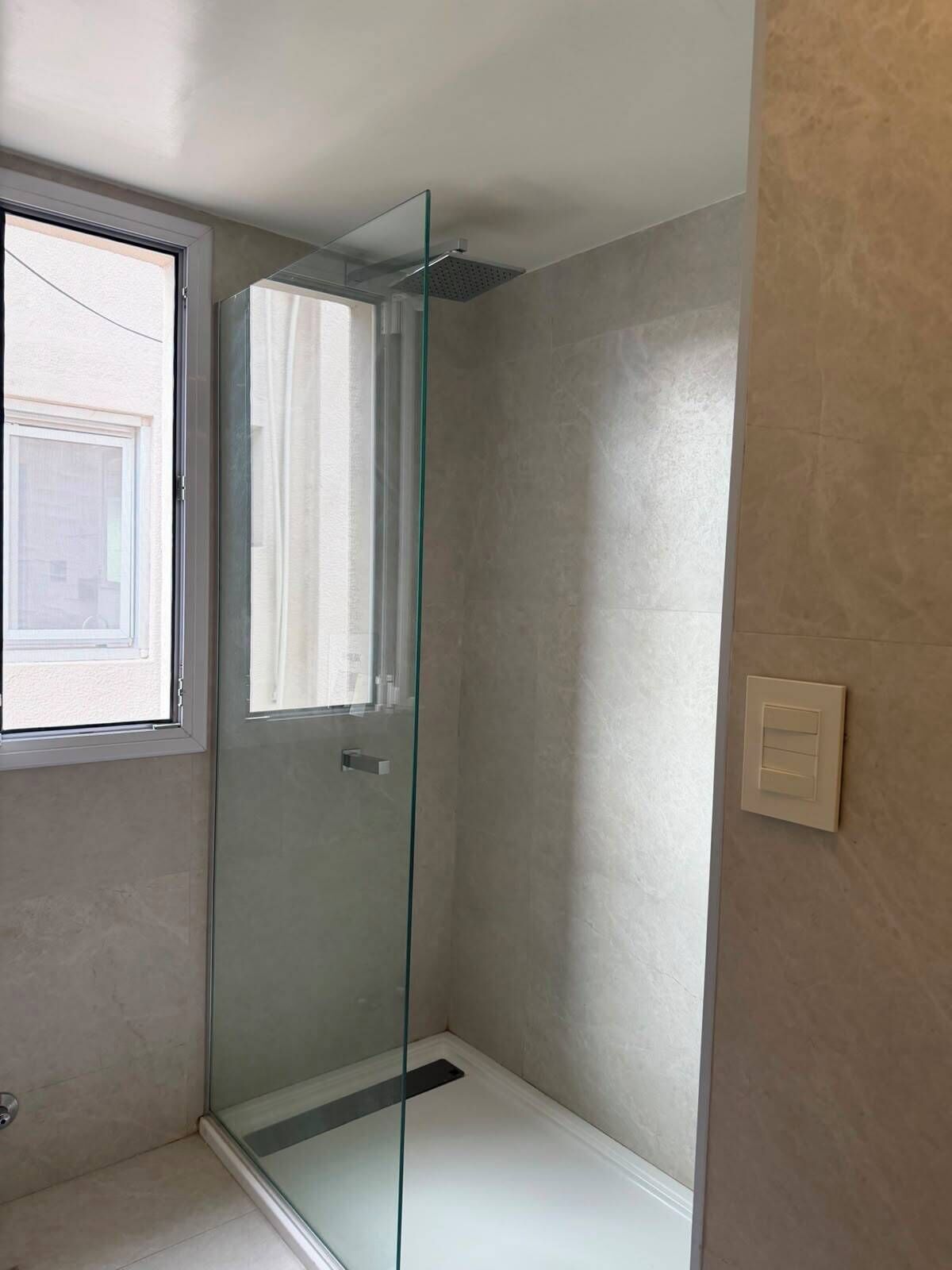 Una ducha con cerramiento de vidrio, una ventana, una puerta e interruptor de luz en la pared.