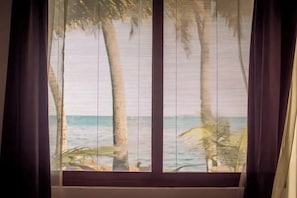 A window with horizontal blinds, a view of palm trees, and a body of water.