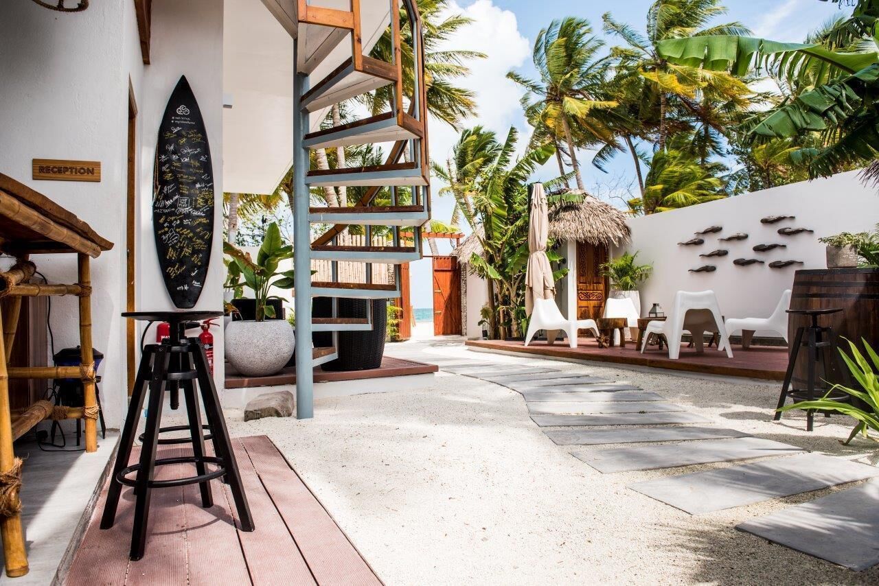 A tropical resort with a reception desk, a surfboard with messages, a staircase, outdoor seating, and palm trees.