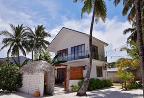 Une maison moderne à deux étages, dotée d’un balcon, entourée de palmiers et agrémentée d’une structure au toit de chaume.