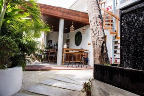 Une cour avec une table et des chaises en bois, une grande plante en pot et un escalier menant à un niveau supérieur.