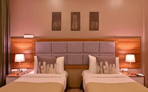 Une chambre d’hôtel avec une tête de lit en bois, deux lits avec des oreillers assortis, des lampes de chevet et des œuvres d’art accrochées au mur.