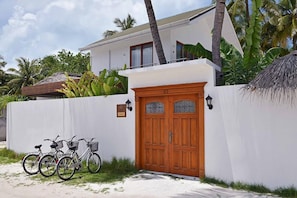 A white building with a wooden door and two windows, surrounded by a white wall and tropical vegetation.
