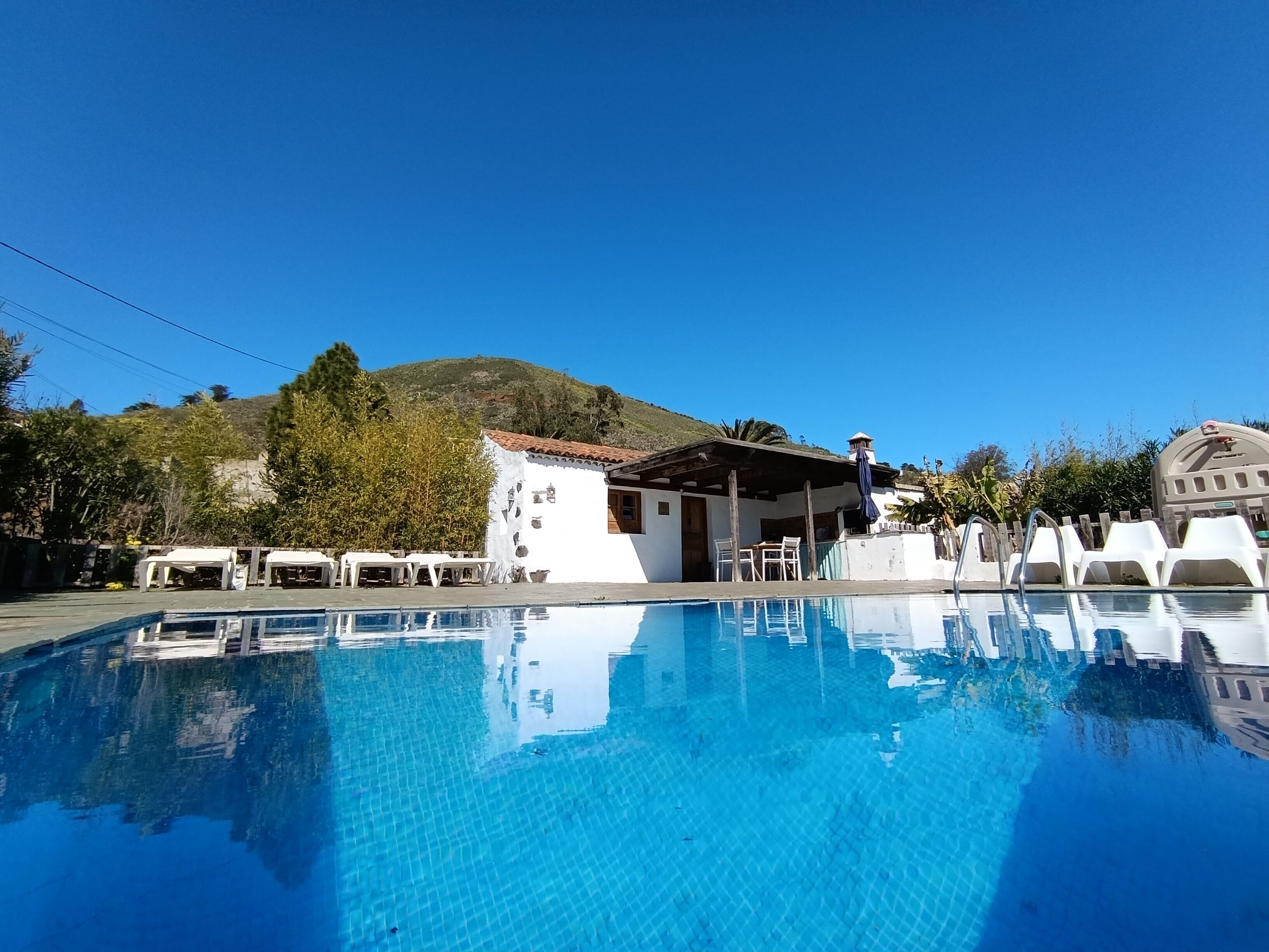 A clear swimming pool with a tiled deck, surrounded by white lounge chairs and a small building with a tiled roof.