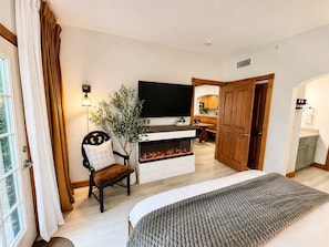 Une chambre à coucher comprenant un lit, une télévision, une cheminée, un fauteuil et donnant sur une cuisine.