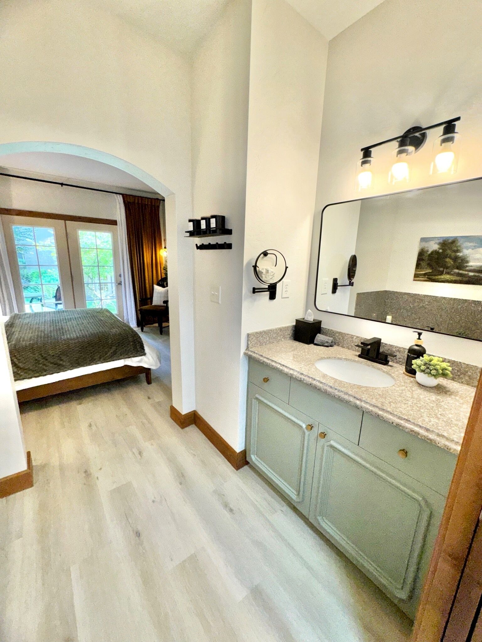 Ein Schlafzimmer mit Bett, ein Badezimmer mit Waschbecken und Spiegel sowie ein Blick ins Freie durch ein Fenster.