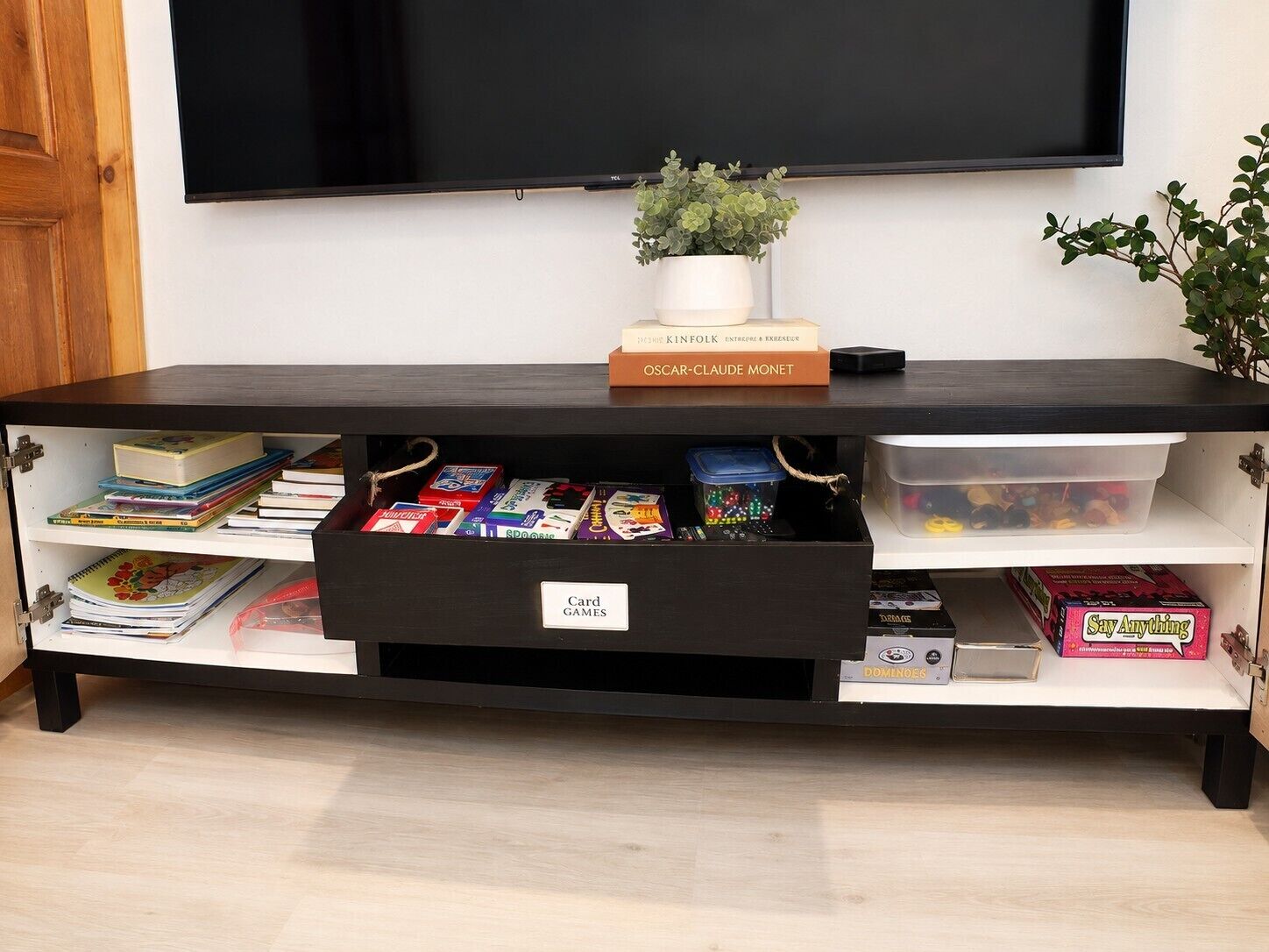 A black entertainment center with a TV mounted above it, a plant on top, and various board games and cards on shelves.