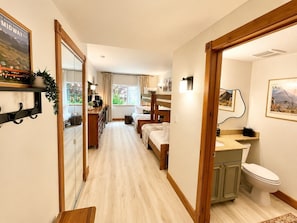 Una camera d'albergo con pavimento in legno, un letto con testiera, una scrivania con sedia, un bagno con toilette e lavandino, e una finestra con tende.