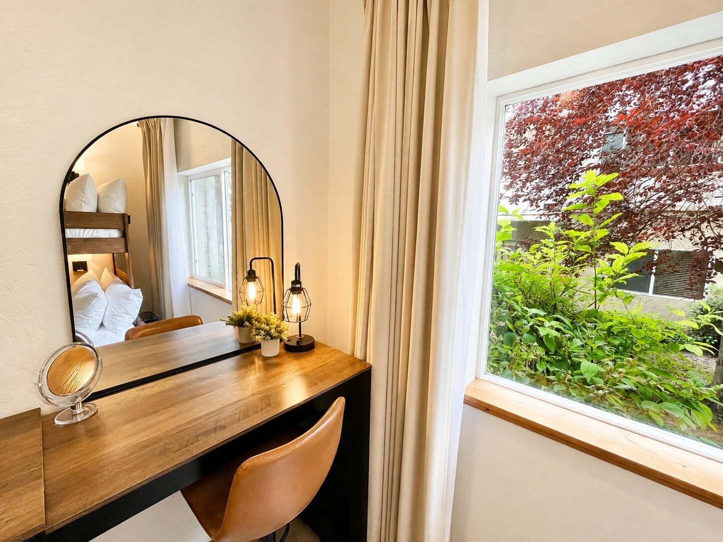Une chambre d’hôtel avec un bureau en bois, un miroir rond, deux lampes fixées au mur, une chaise et une fenêtre donnant sur un paysage verdoyant avec un arbre.