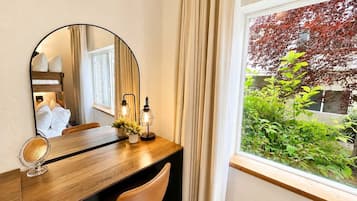 A hotel room with a wooden desk, a round mirror, two wall-mounted lights, a chair, and a window with a view of greenery and a tree.
