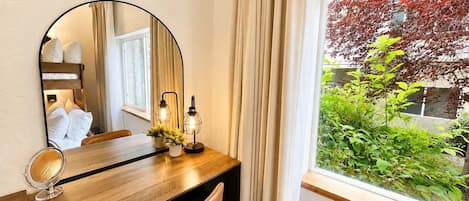 Ein Hotelzimmer mit einem Holztisch, einem runden Spiegel, zwei Wandleuchten, einem Stuhl und einem Fenster mit Blick auf Grünflächen und einen Baum.