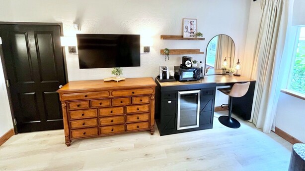 A room with a wooden dresser, a desk with a computer, a flat-screen TV mounted on the wall, and a mirror.