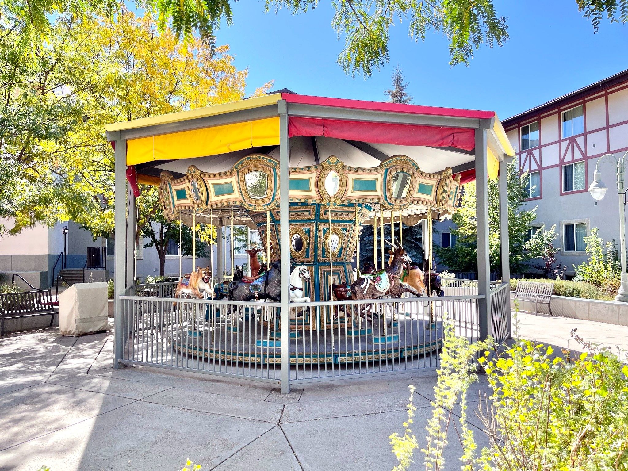 Un carrousel aux décorations dorées et travaillées, surmonté d’un toit rouge, entouré d’une clôture métallique, et installé dans un parc agrémenté de bancs et d’arbres.