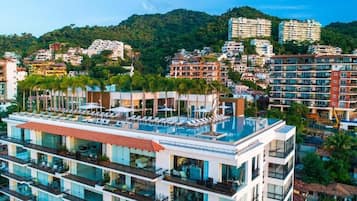 Un hotel de varios pisos con un área de piscina en la azotea, rodeado de otros edificios y un paisaje montañoso.