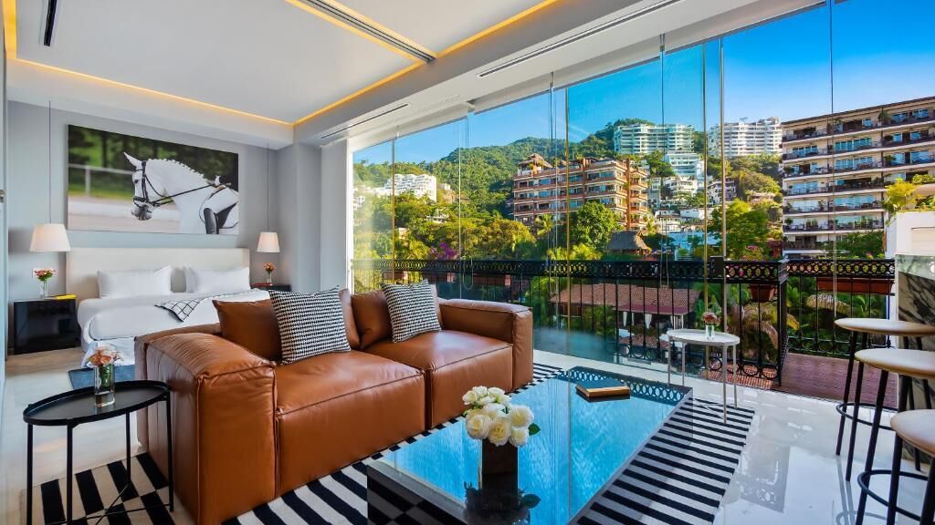 A modern hotel room with a large window, a brown leather sofa, a glass coffee table, and a view of a cityscape.