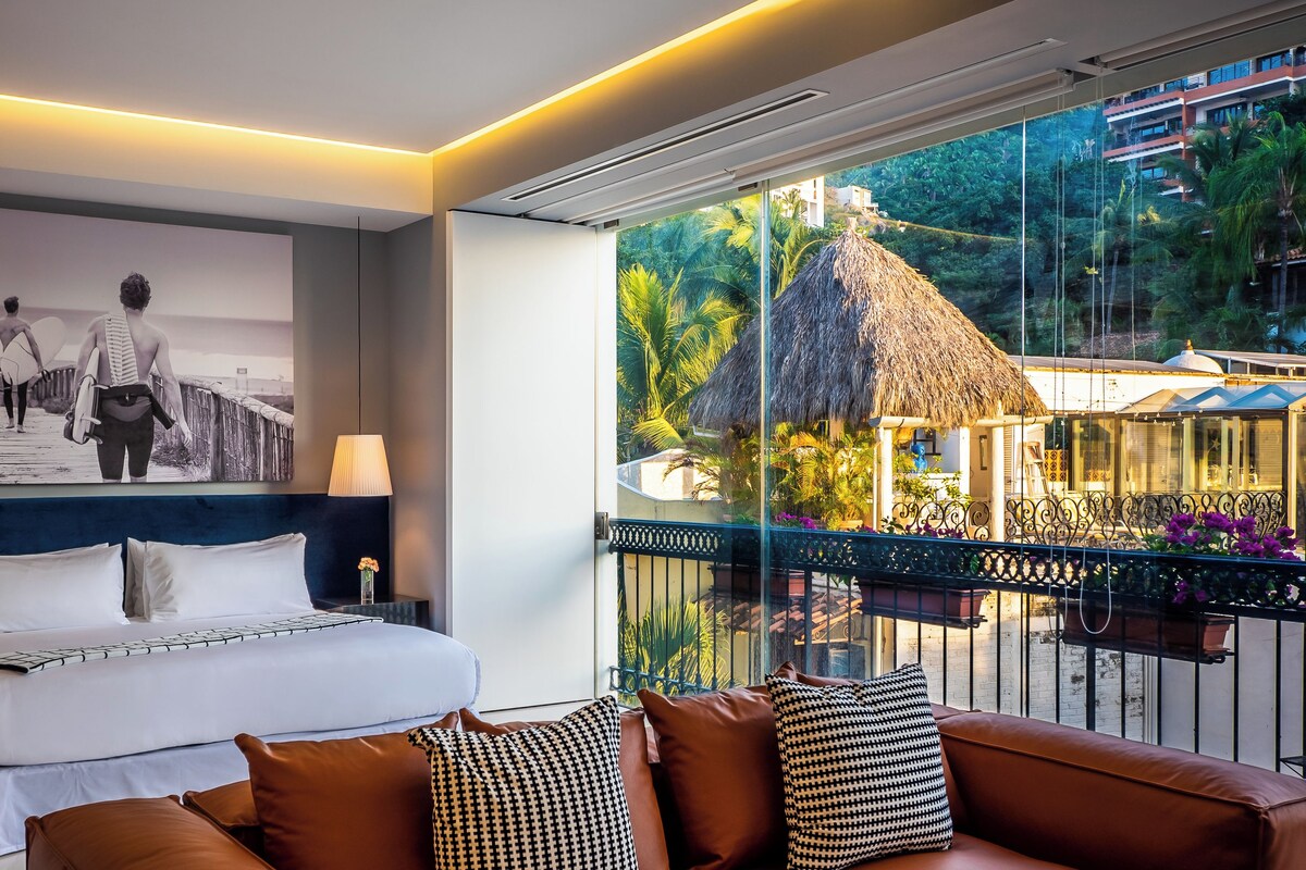 Ein modernes Hotelzimmer mit einem Bett, einem Sofa, einem großen Fenster mit Blick auf ein Gebäude mit einem Reetdach und einem gerahmten Bild an der Wand.