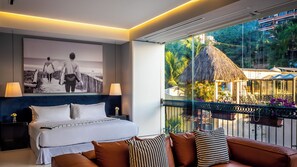 Una habitación de hotel moderna con una cama, un sofá, un ventanal con vistas a una estructura de techo de paja y un cuadro enmarcado en la pared.