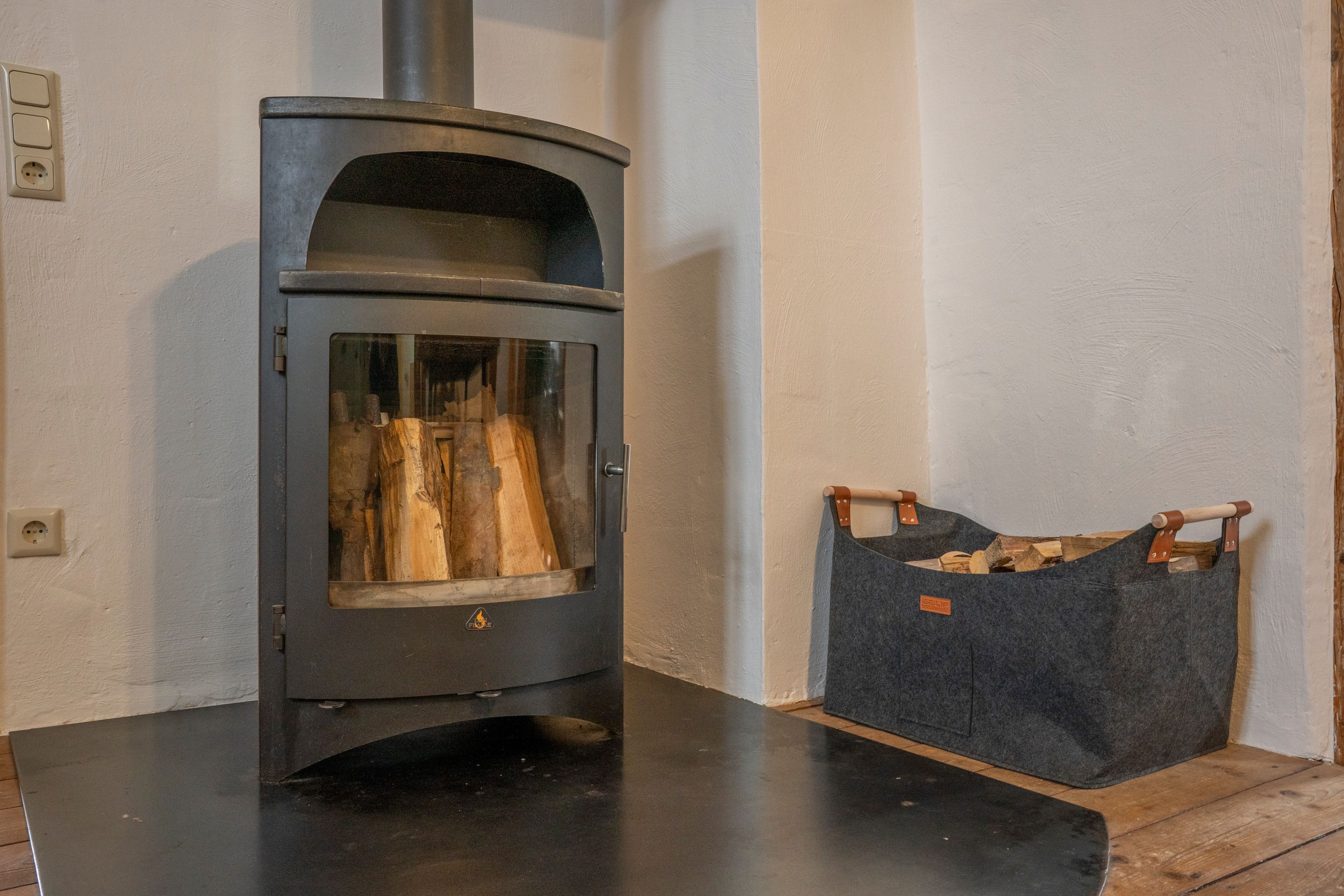 Een houtkachel met glazen deur, een mand voor hout en een stopcontact op de muur.