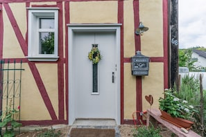 Una casa tradicional con entramado de madera y techo de paja, con una puerta blanca, una ventana pequeña y un cartel de bienvenida.