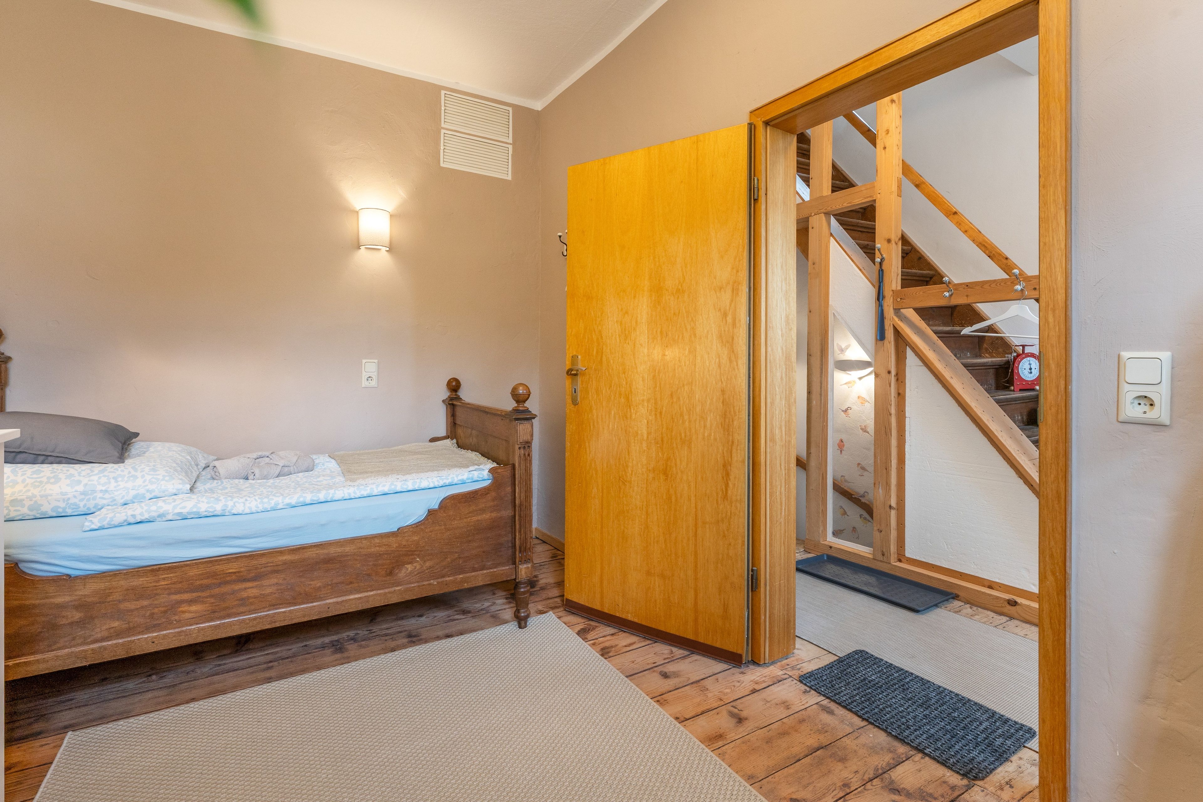 Een slaapkamer met een houten bed, een deur naar een trap en een wandlamp.