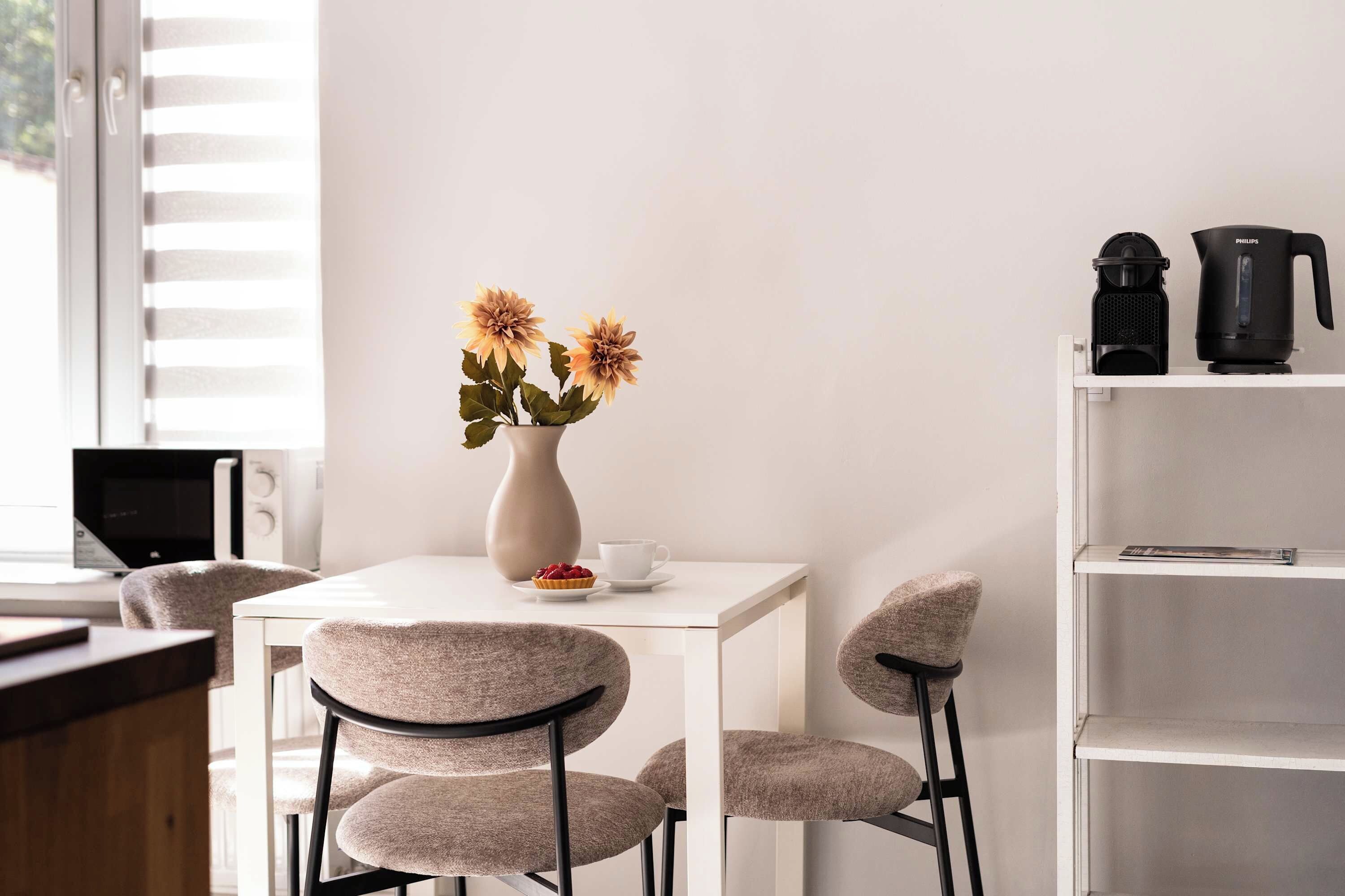A modern room with a white table, two chairs, a vase with flowers, a coffee maker, and a TV.