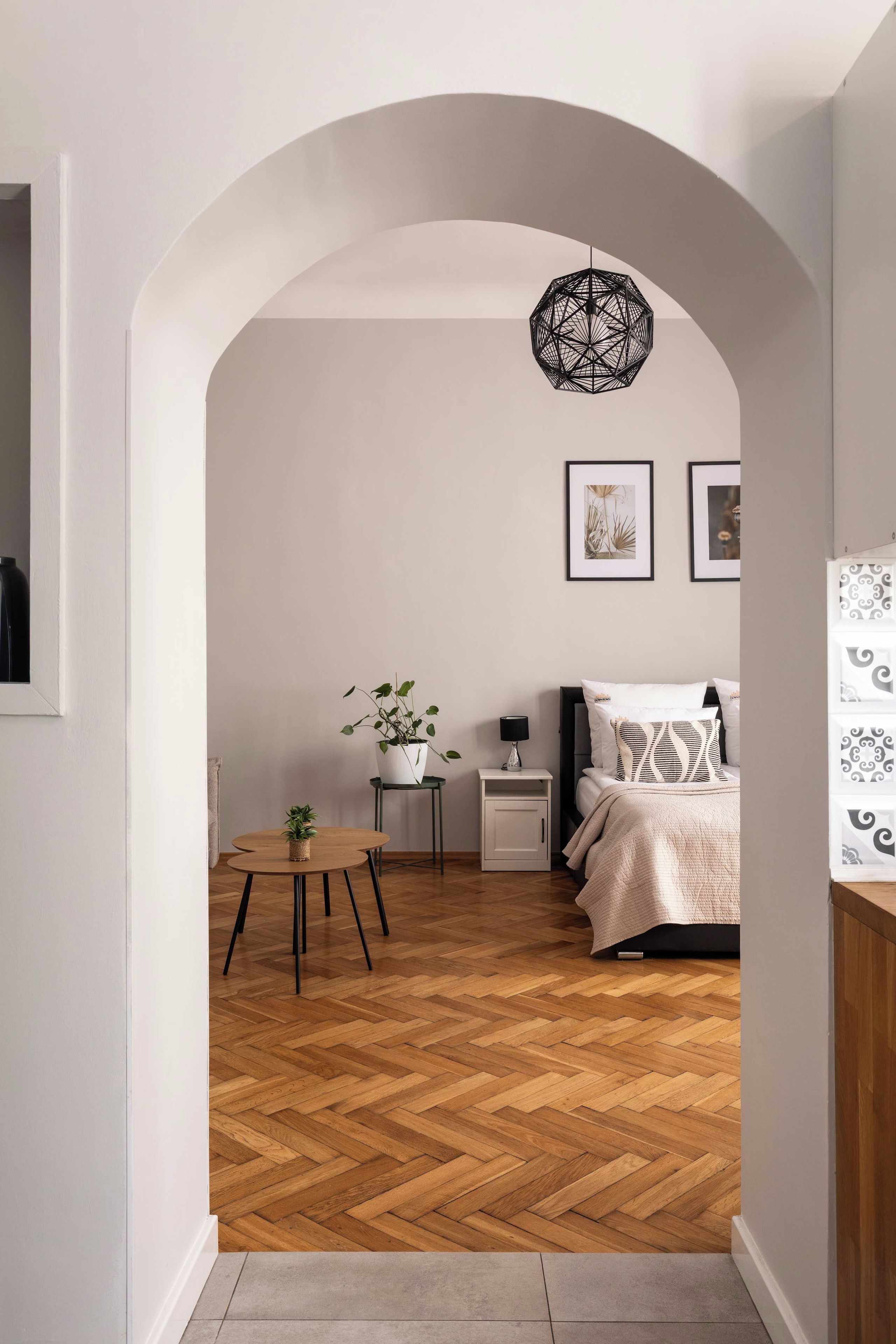 Um corredor leva a um quarto com piso em herringbone, uma mesa de madeira redonda, um vaso de plantas, um abajur e uma cama com manta estampada.