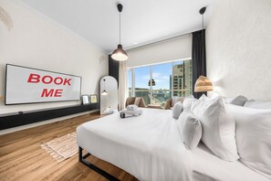 Una camera d'albergo moderna con un letto grande, vista sulla città, un cartello con scritto 'BOOK ME' e un'ampia finestra.