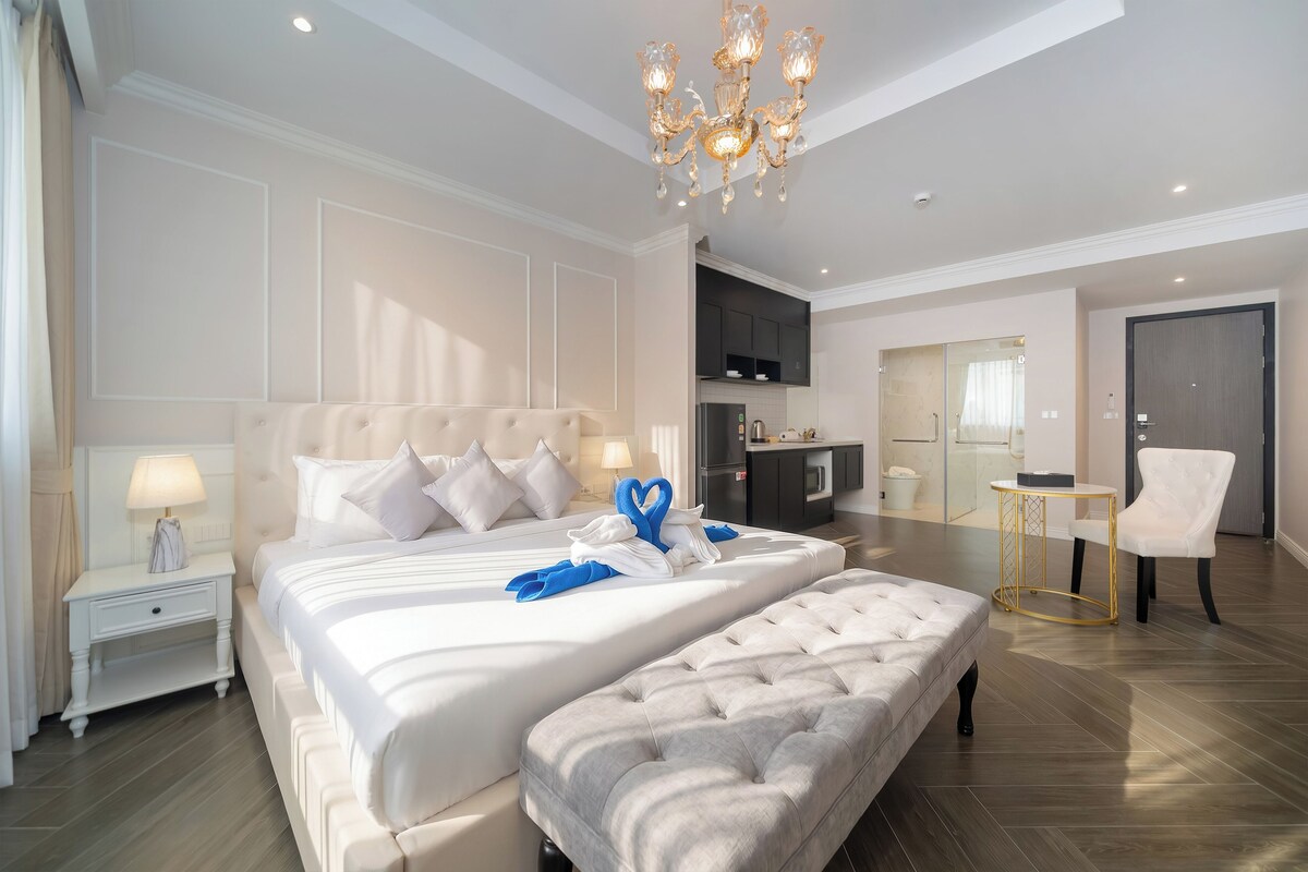 A hotel room with a large bed, a bench, a desk, a chair, a kitchenette, and a bathroom.