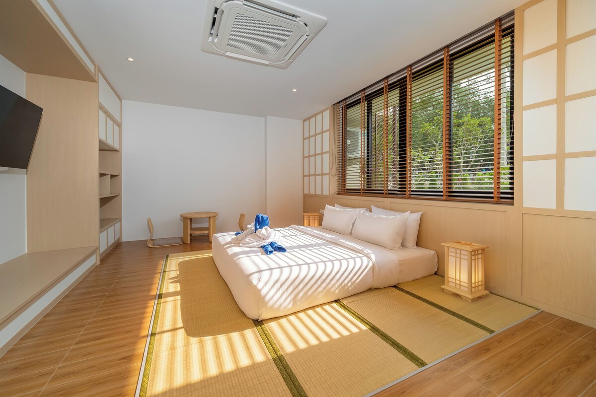 A modern bedroom with a large bed, a bedside table, a small stool, and a window with blinds.