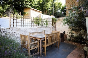 A wooden outdoor seating area with a table and benches, surrounded by a white brick wall and greenery. - A Battersea State of Mind (London)