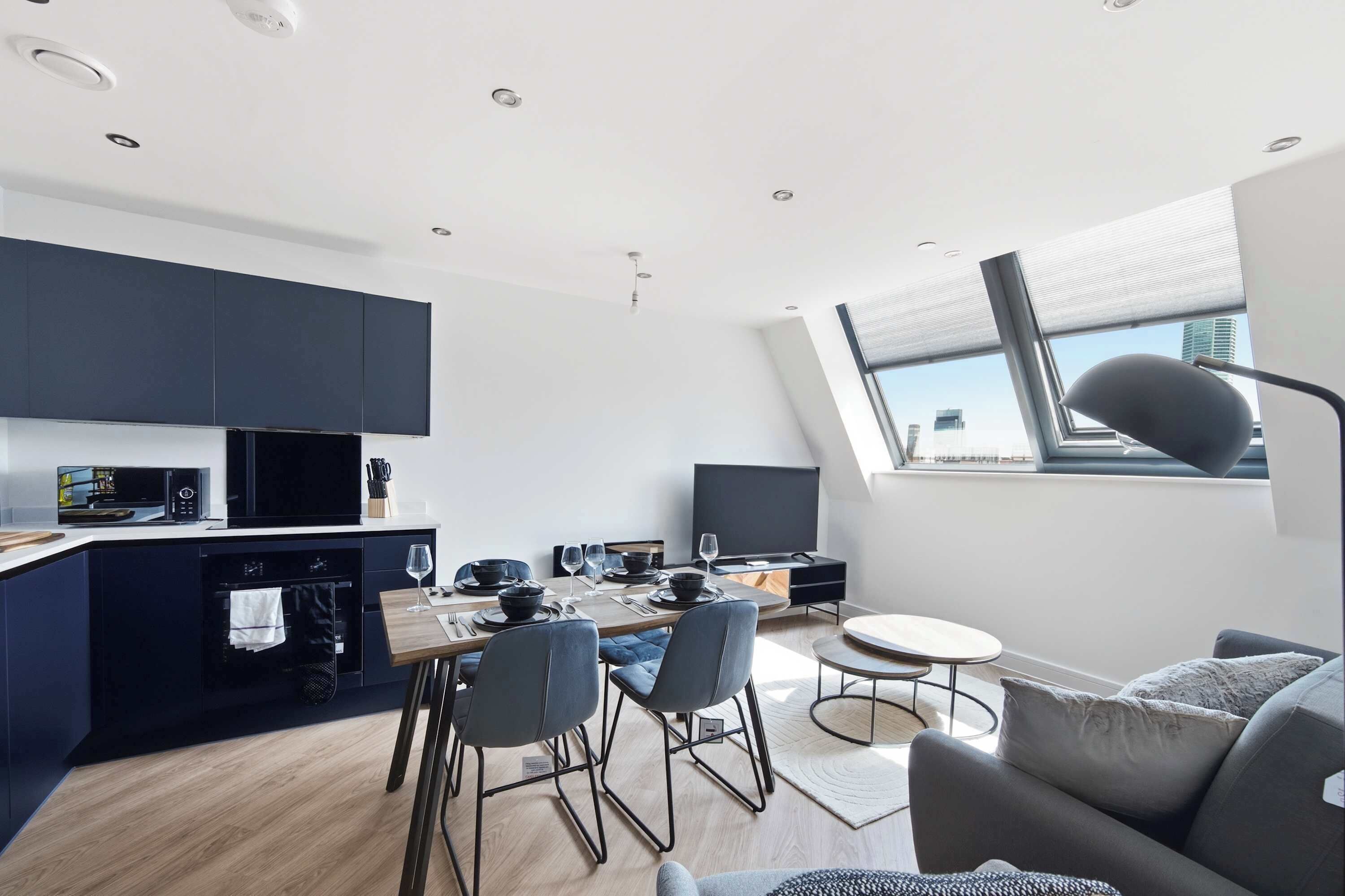 Eine moderne Küche mit dunklen Schränken, ein Essbereich mit einem Tisch für vier Personen und ein großes Fenster mit Blick auf die Stadt.