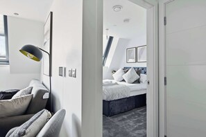 A modern hotel room with a grey sofa, a black floor lamp, and a bed with a grey headboard. - Modern 1 Bedroom Apartment in Central Birmingham (Birmingham)