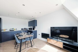 Eine moderne Küche mit dunklen Schränken, ein Essbereich mit einem Holztisch und blauen Stühlen sowie ein großer Flachbildfernseher an der Wand.