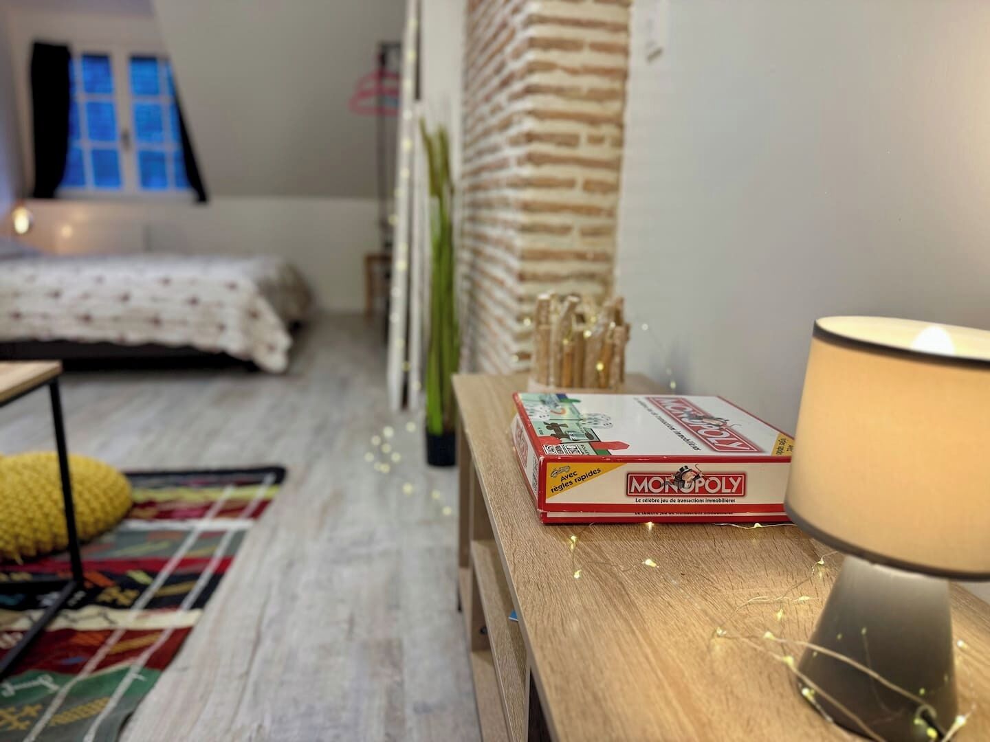 Une pièce confortable avec un lit, une table en bois sur laquelle est posé un jeu de société Monopoly, une lampe et un tapis à motifs.