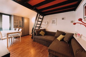 Uma sala de estar acolhedora com piso de madeira, um sofá marrom, uma mesa branca com cadeiras e uma área de cozinha com armários e uma escada.