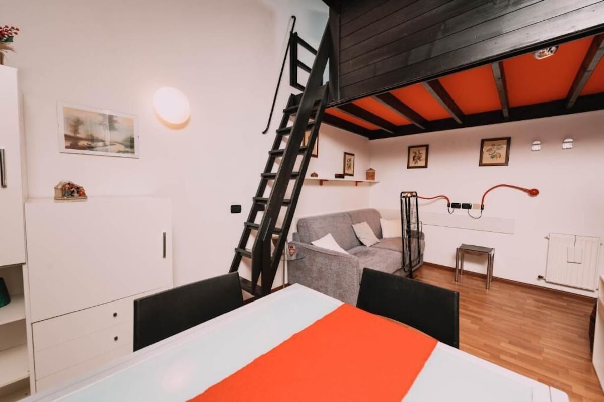 Quarto com uma mesa de jantar, um sofá cinza, uma escada e teto de madeira.