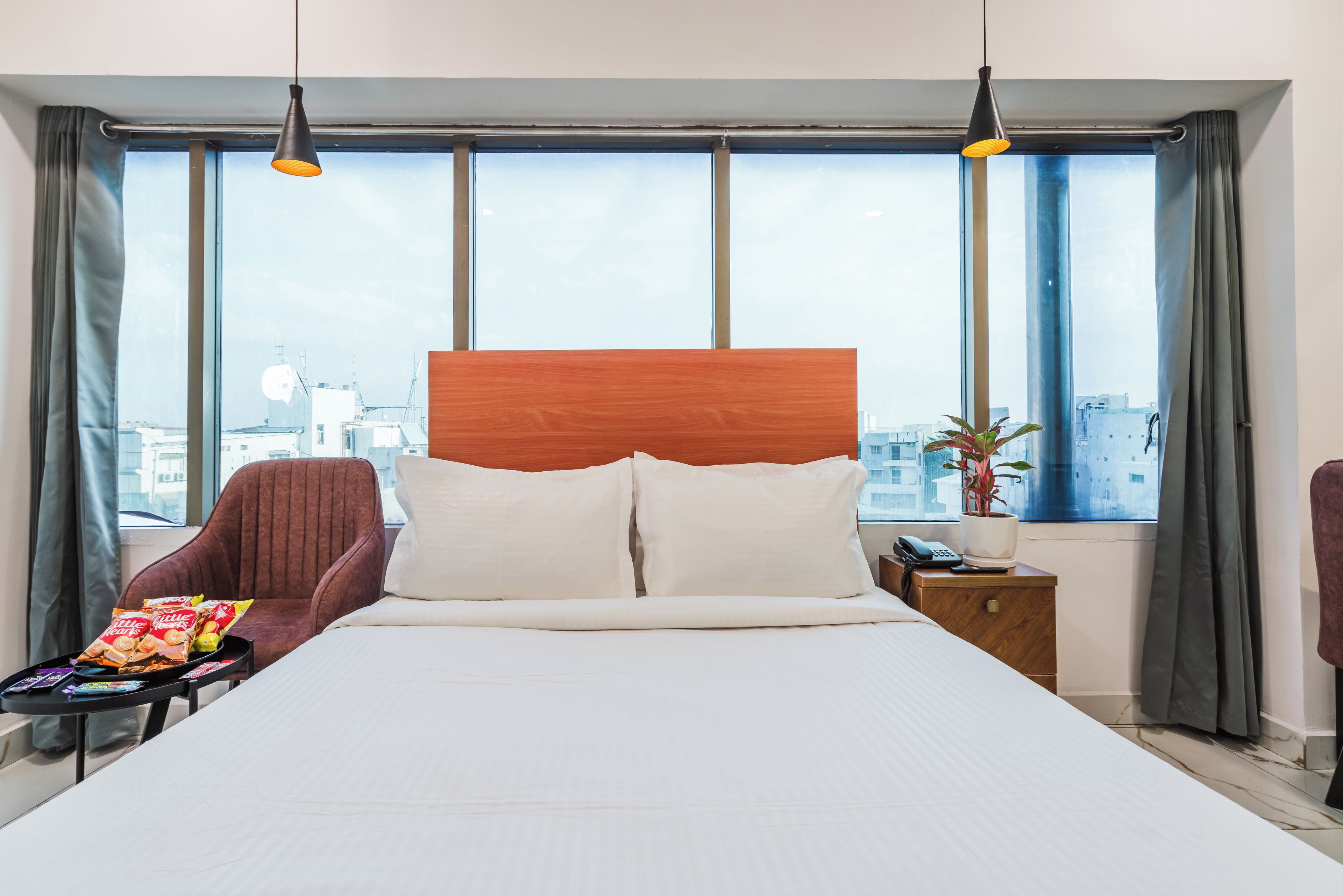 A hotel room with a large bed, a wooden headboard, two bedside tables with lamps, a chair, a small table with snacks, and a view of the city through large windows.