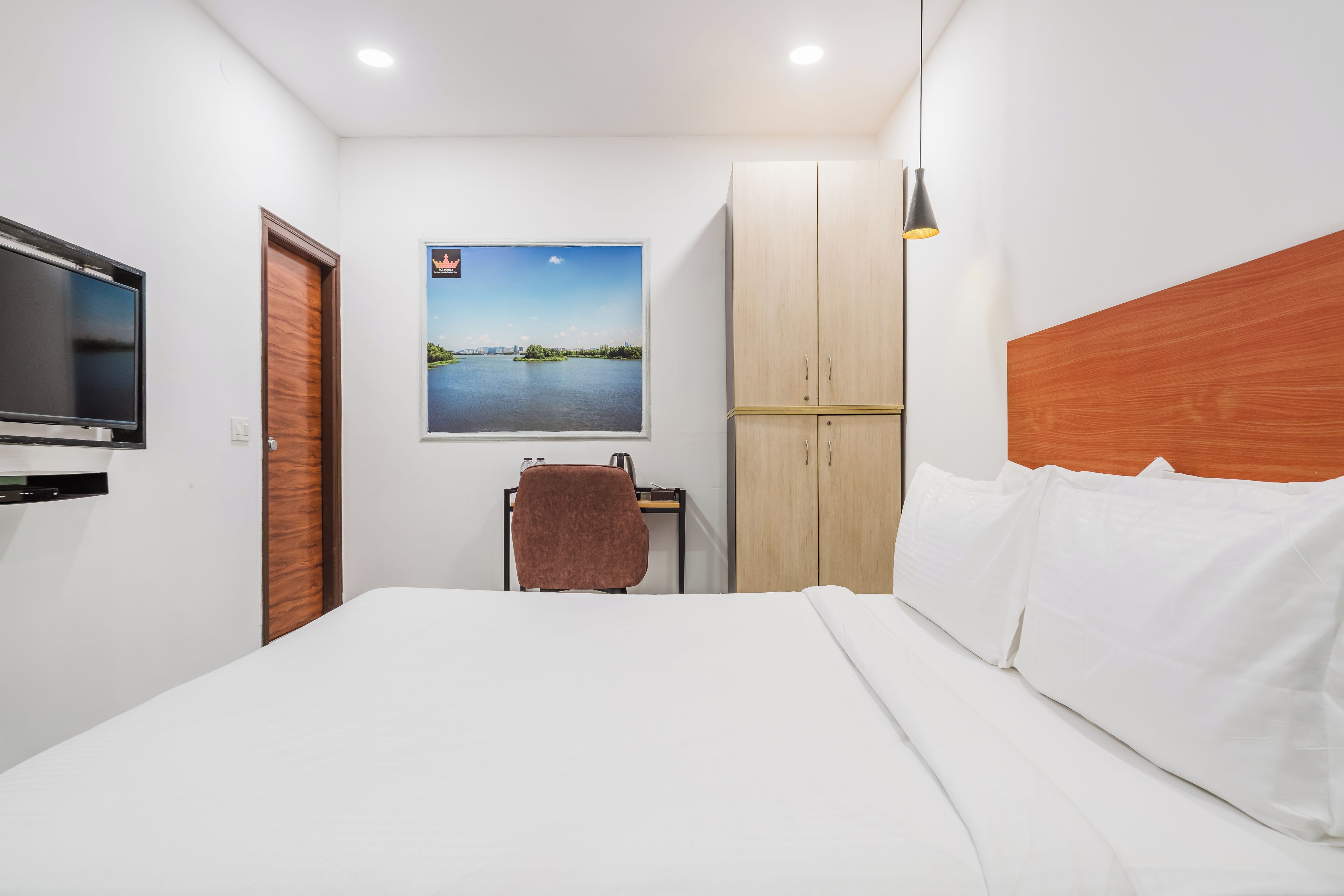 Une chambre d’hôtel comprenant un lit, une télévision, une armoire en bois, un bureau avec une chaise et un tableau représentant un paysage.