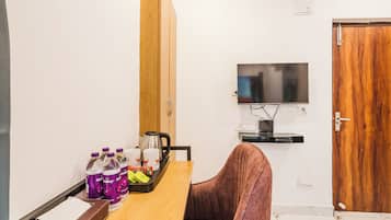 Habitación pequeña con puerta de madera, televisor montado en la pared, mesa con silla y bandeja con agua embotellada y vasos.