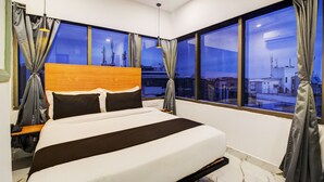 Quarto de hotel moderno com cama grande, cabeceira de madeira e vista para a cidade através de amplas janelas.