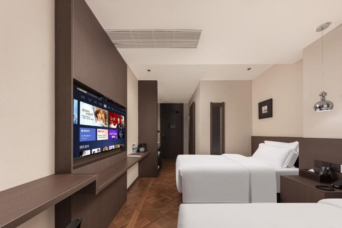 A hotel room with a TV mounted on the wall, a bed, a bedside table, and a desk with a chair.