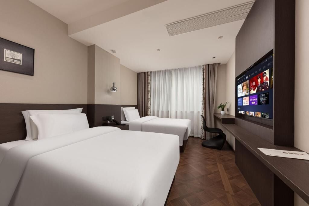 Habitación de hotel con dos camas, un televisor mostrando una película, un escritorio con una silla y una ventana con cortinas.