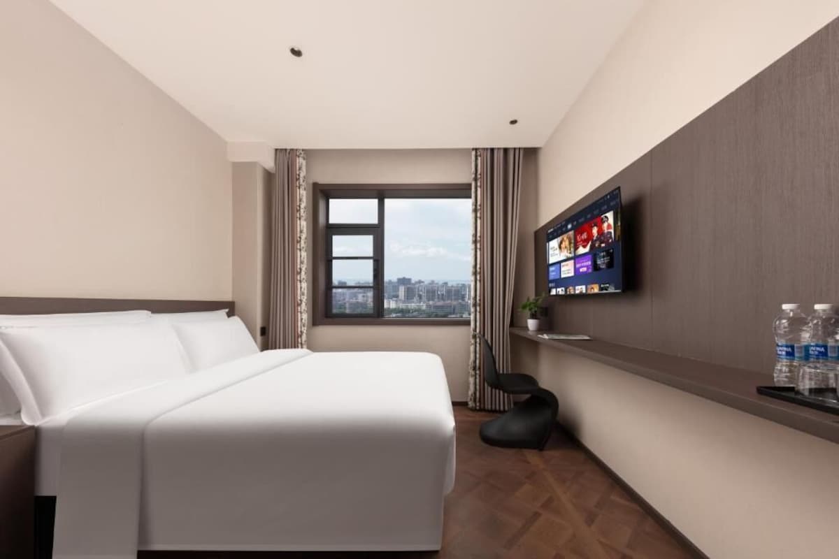 A hotel room with a bed, a TV mounted on the wall, a window with a city view, and a small shelf with water bottles.
