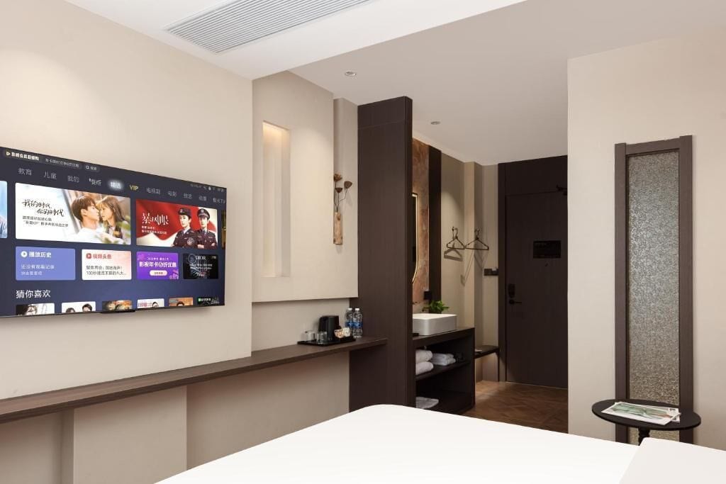 Habitación de hotel con un televisor de pantalla plana grande montado en la pared mostrando varios contenidos. Cuenta con una cama con cabecera blanca, una mesita de noche con lámpara y un estante con artículos de aseo.