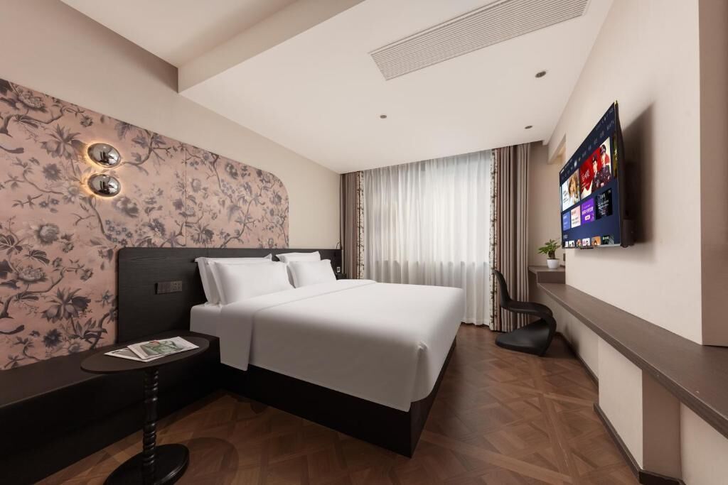 A hotel room with a large bed, a flat-screen TV mounted on the wall, a small table with a magazine, and a decorative wall with floral patterns.