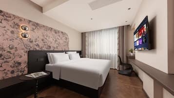 A hotel room with a large bed, a flat-screen TV mounted on the wall, a small table with a magazine, and a decorative wall with floral patterns.