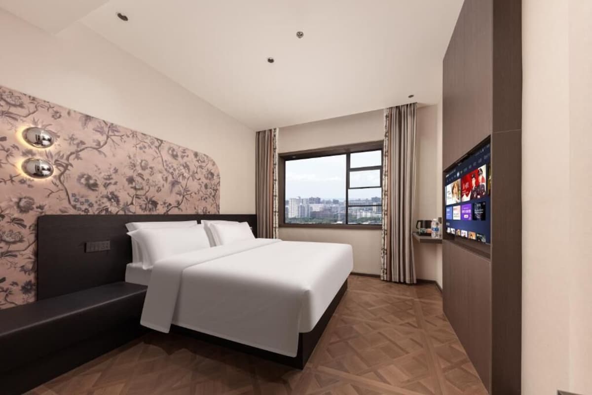 A hotel room with a bed, a seating area, a television, and a window with a city view.