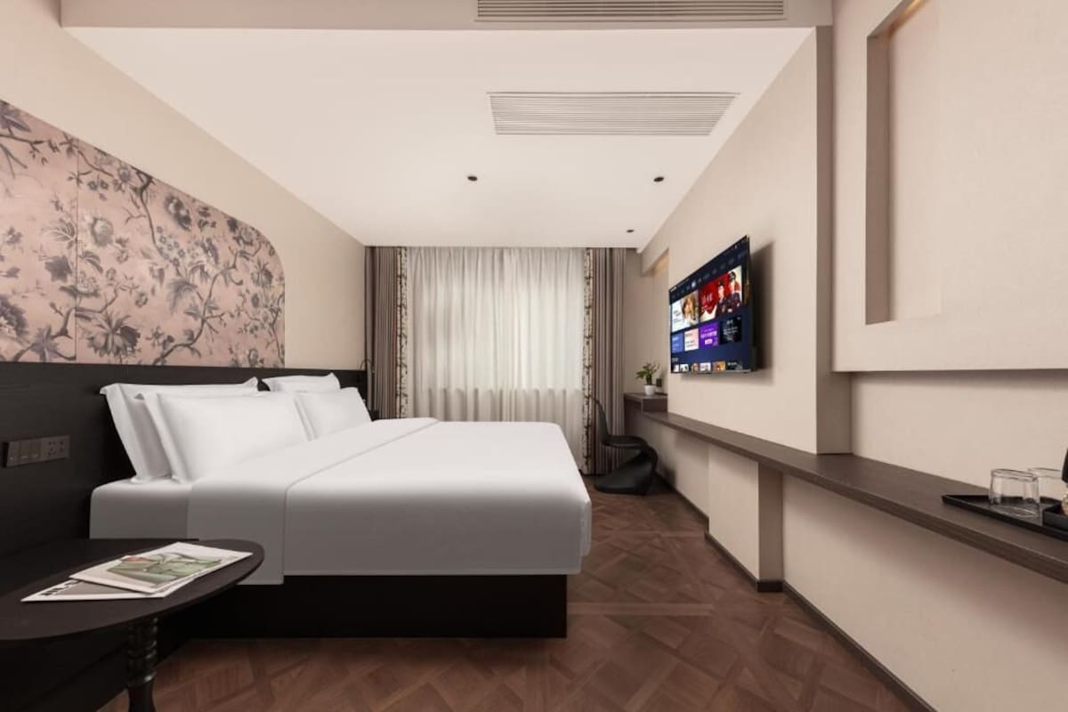 A modern hotel room with a bed, a TV mounted on the wall, a small table with a magazine, and a wooden floor.