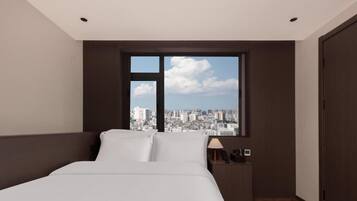 A hotel room with a bed, a nightstand, a lamp, and a window with a city view.